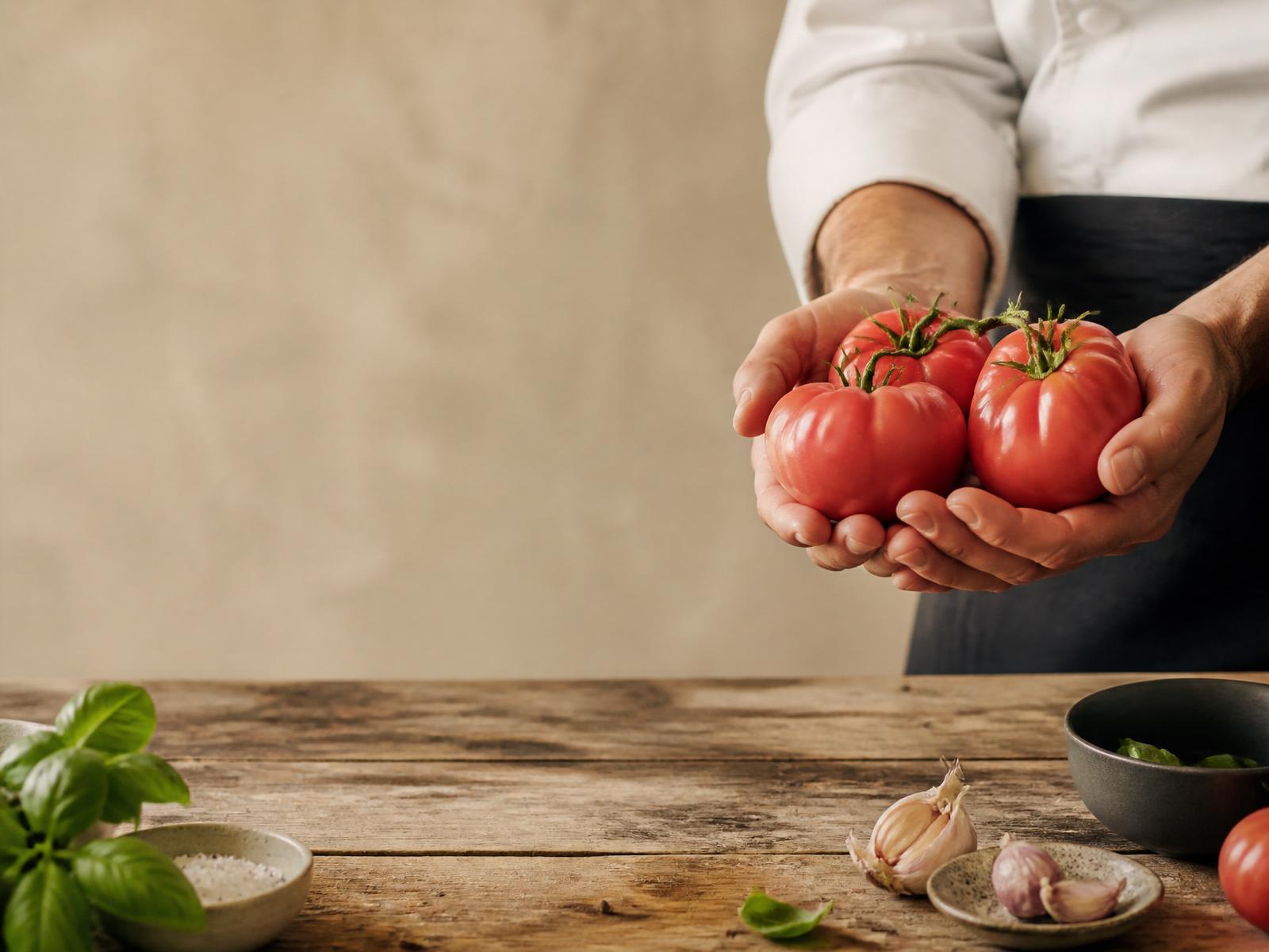 A chef holding fresh ripe tomatoes over a rustic kitchen table